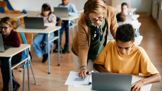 A teacher assists her elementary student in using a laptop in class.