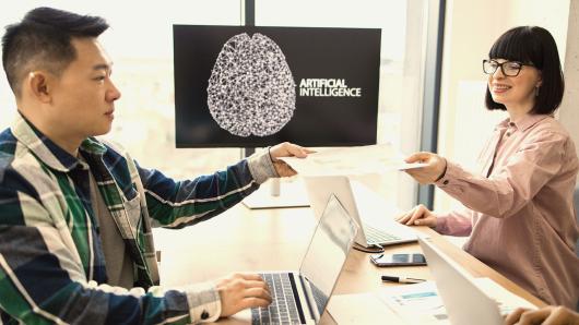 Two smiling business professionals sit in front of laptops. A computer screen behind them reads "artificial intelligence".