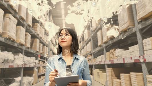 A smiling young woman looks at inventory in warehouse while using a smart tablet.