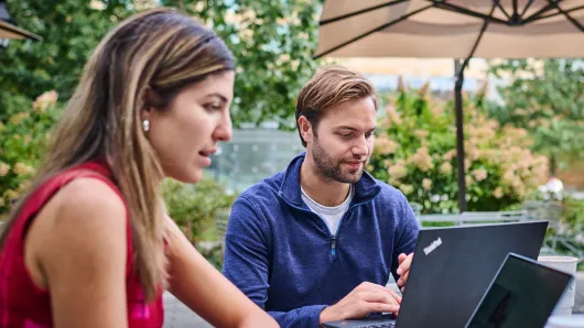 A female and a male participant sit outside at a table together while separately working on their laptops