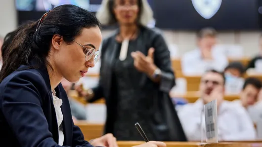 A female participant takes notes inside the classroom as the professor lectures in the background