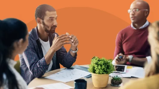 Group of professionals in a meeting, engaged in discussion around a table with papers, a tablet, coffee mugs, and small plants, set against an orange background.