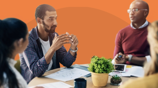 Group of professionals in a meeting, engaged in discussion around a table with papers, a tablet, coffee mugs, and small plants, set against an orange background.