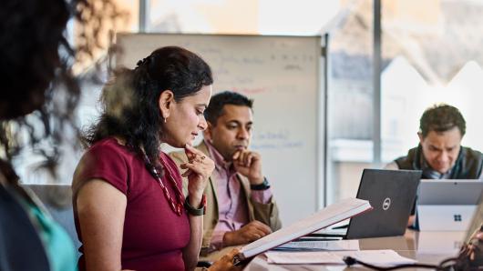 Male and female executives review their notes on paper and on their laptops while sitting down inside a meeting room with a white board in the background