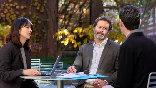 Three professionals meet around an outdoor table with a laptop and documents in a campus courtyard.