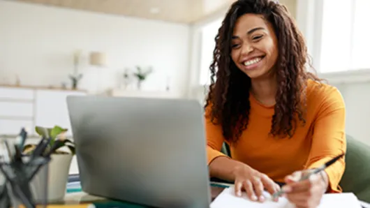 Image of a woman in an orange shirt sitting with a computer writing something on a note pad.