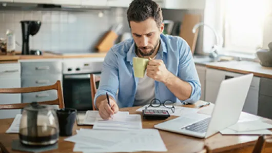 Man sitting at a kitchen table reviewing paperwork while holding a mug, with a laptop and calculator beside him.