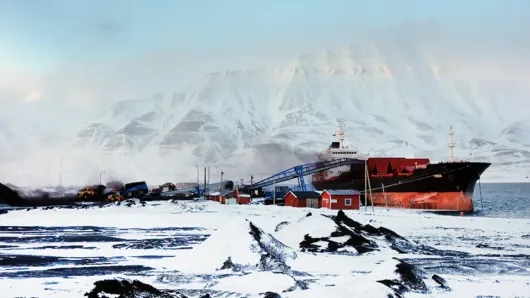 Large cargo ship docked at an industrial port surrounded by snow-covered terrain, with towering Arctic mountains in the background.