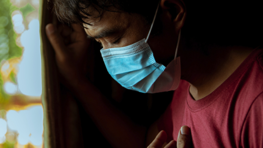 Man wearing a surgical mask holding his chest with eyes closed. 