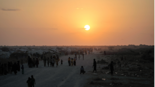 Groups of people in a refugee camp walking beneath an orange sunset sky