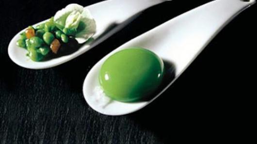 Two spoons, one holding intact peas and a pea flower, the other with a spherification of pea soup.