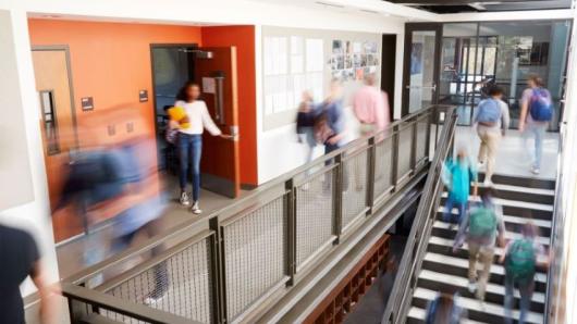 Students and staff walking through a hallway with staircase leading down. Students and staff are blurred to indicate movement.